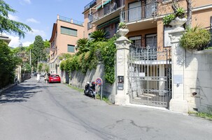 A street with a gated entrance, a red car parked on the side, and a building with balconies and greenery.