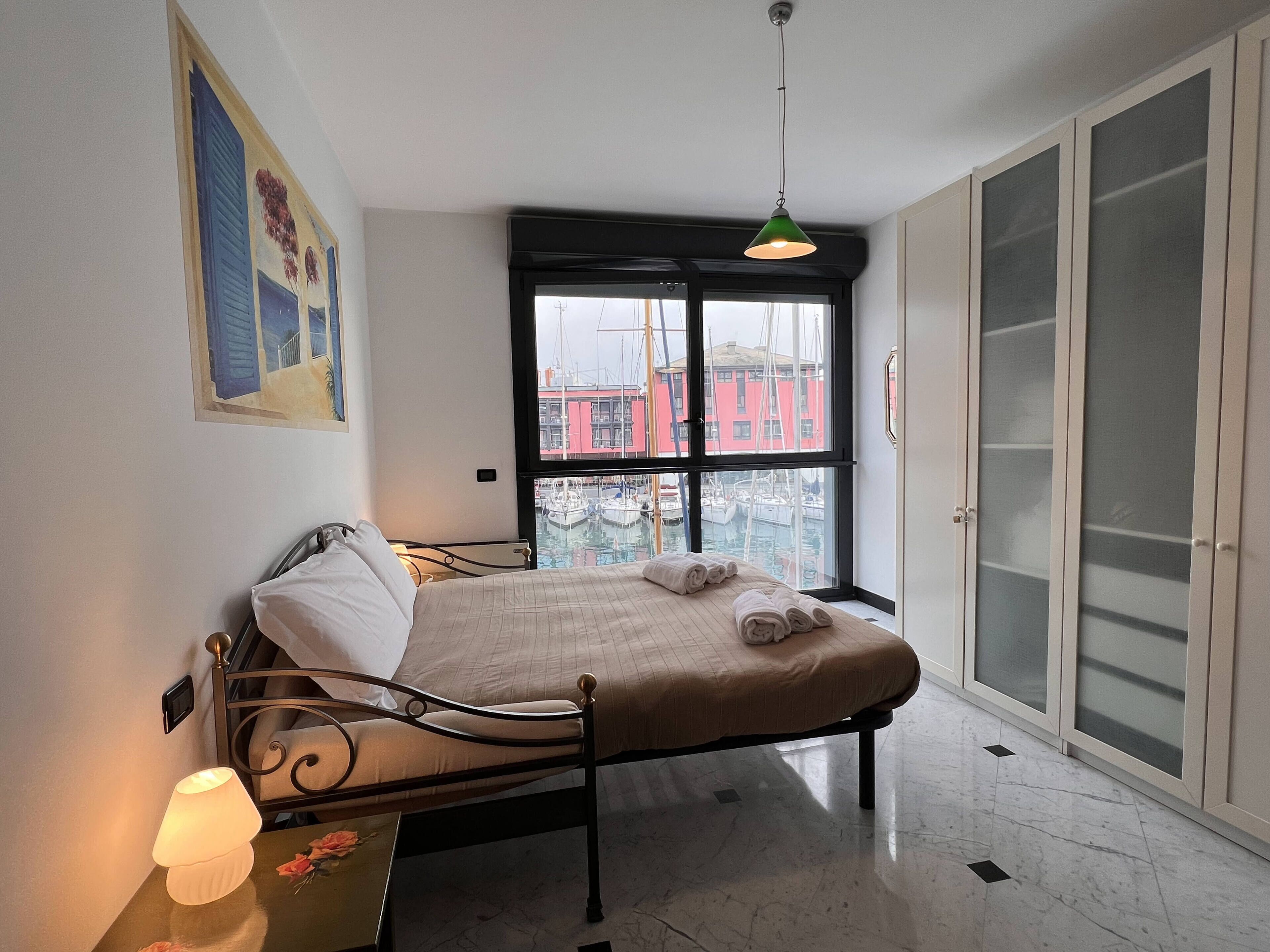 A bedroom with a bed, a bedside table with a lamp, a window with a view of buildings and boats, and a door with frosted glass panels.