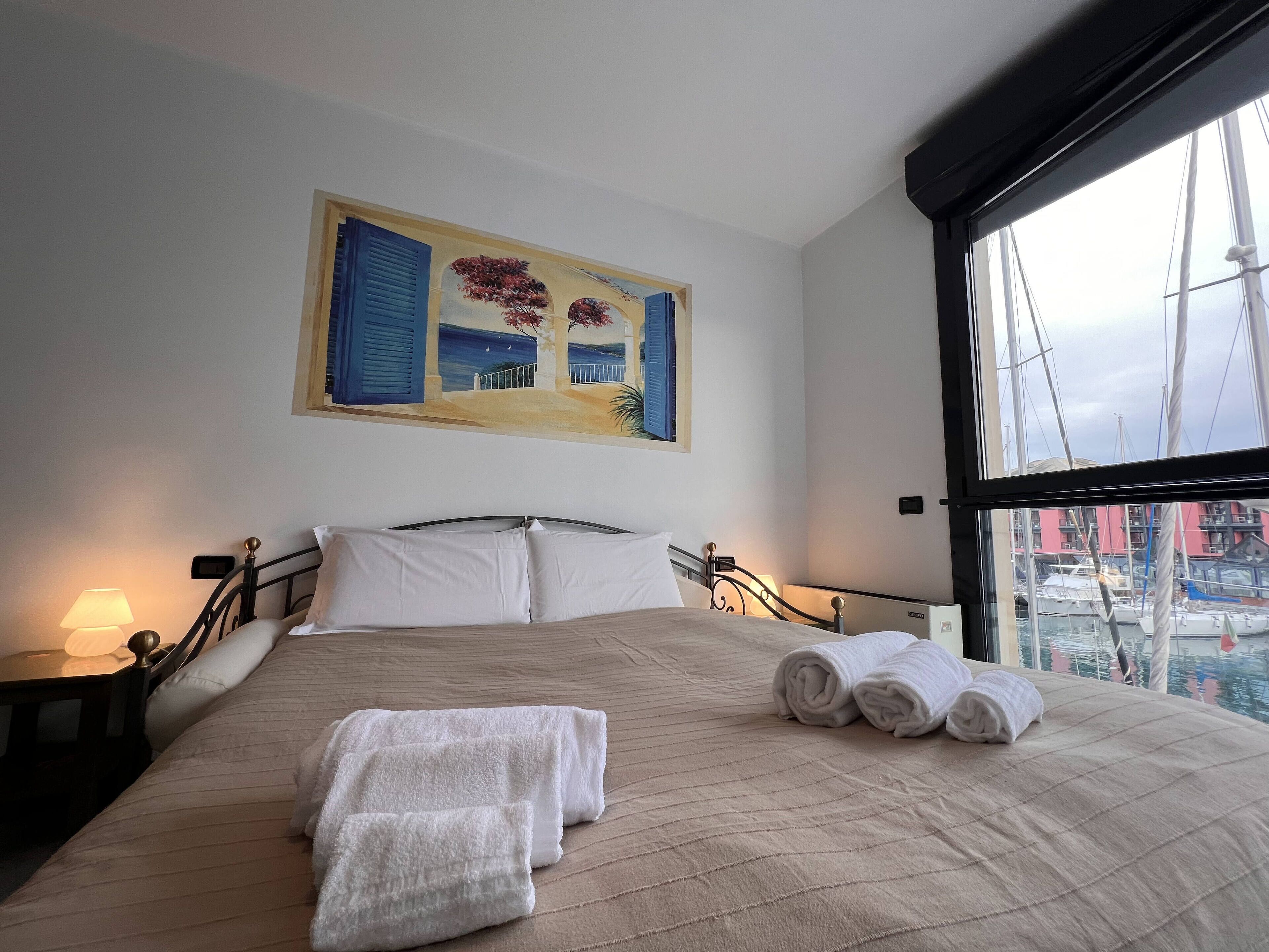 A hotel room with a bed, a painting, a window, and a view of boats.