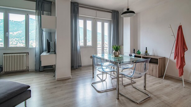 A modern dining area with a glass table and metal chairs, a view of the outdoors through large windows, and a radiator on the floor. - JOIVY Family Flat W/Balcony (Como)