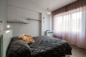 Une chambre à coucher comprenant un lit, une table de chevet, une commode, une étagère avec des objets décoratifs et une fenêtre avec des rideaux.