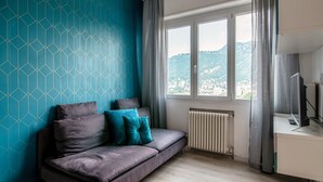 A modern room with a sofa, a desk, and a large window with a view of a city and hills. - JOIVY Family Flat W/Balcony (Como)