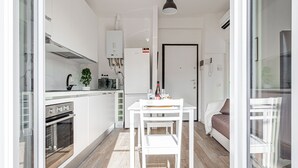A modern kitchen with white cabinets, a built-in oven, a dining table with a chair, and a sofa in the adjacent living area.