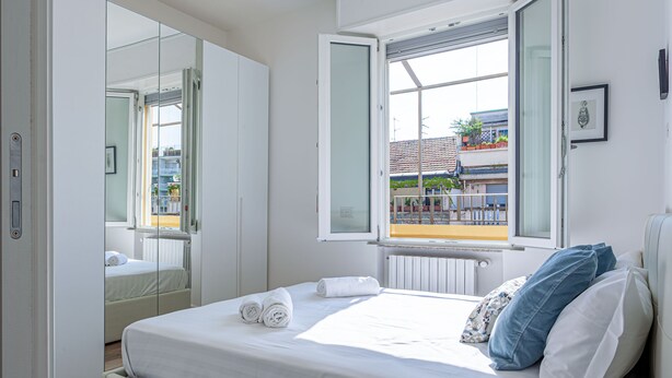 A modern bedroom with a large bed, a window with an open view, a mirror, and a door.