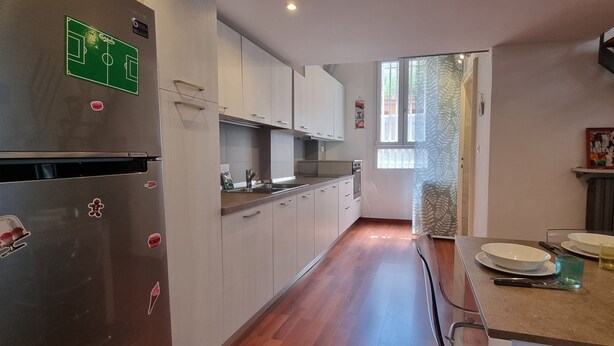A modern kitchen with a stainless steel refrigerator, white cabinets, a wooden dining table with plates and glasses, and a staircase leading to another level. - JOIVY Studio Near Porta Romana (Milan)