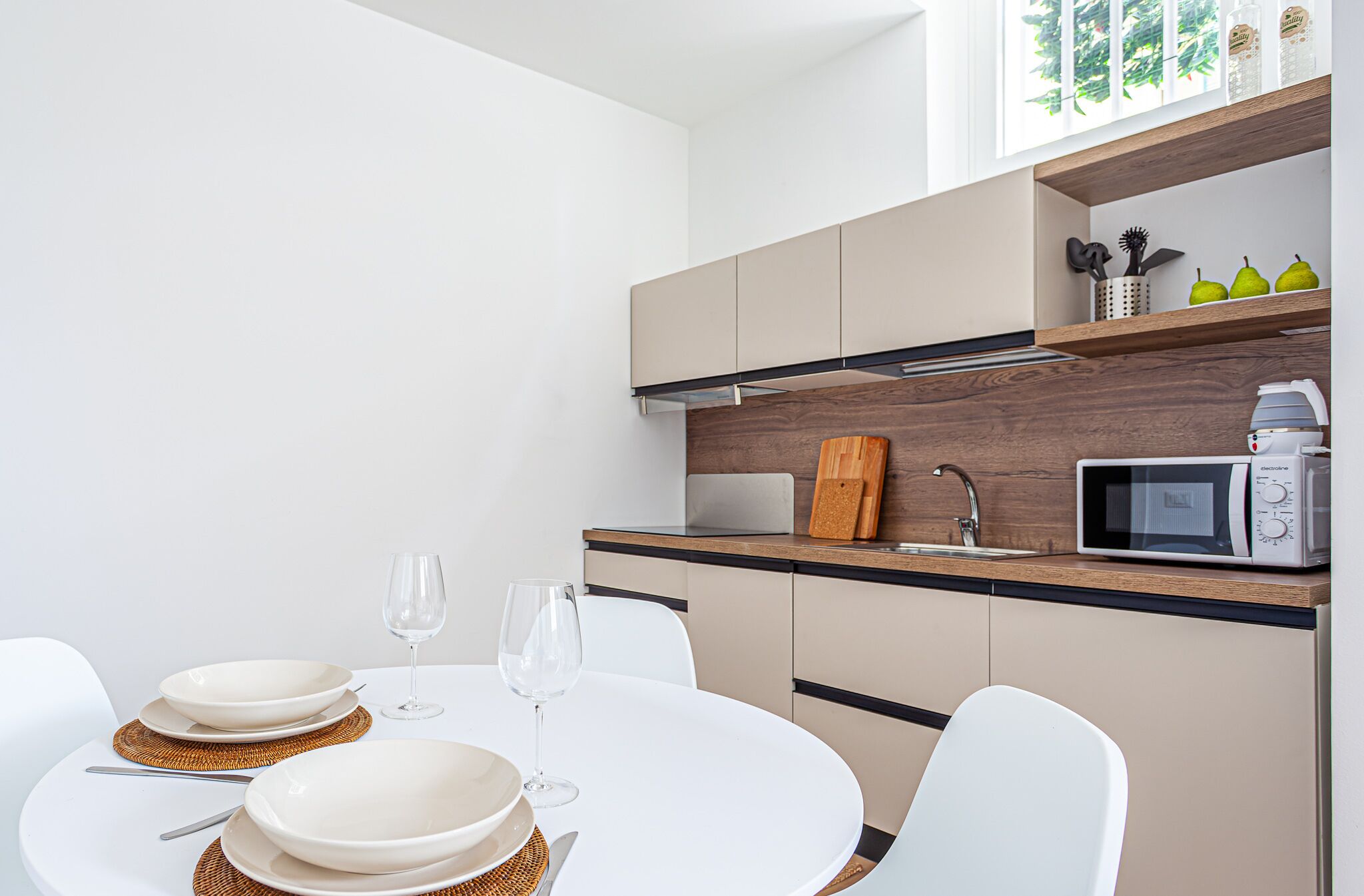A modern kitchen with white cabinets, a microwave, and a wooden countertop. There is a dining table with white chairs and a wine glass.
