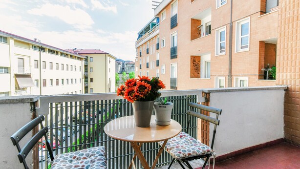 Un balcón con una mesa redonda y dos sillas, una planta en maceta y vistas de edificios circundantes.