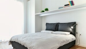 Un dormitorio moderno con una cama, cabecera blanca, repisa con libros y plantas, y suelo de madera.