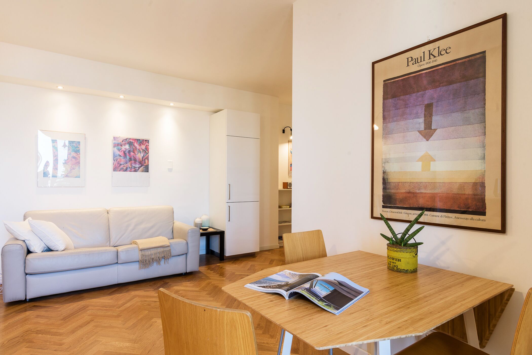 A modern living room with a white sofa, wooden dining table, and a framed artwork of Paul Klee.