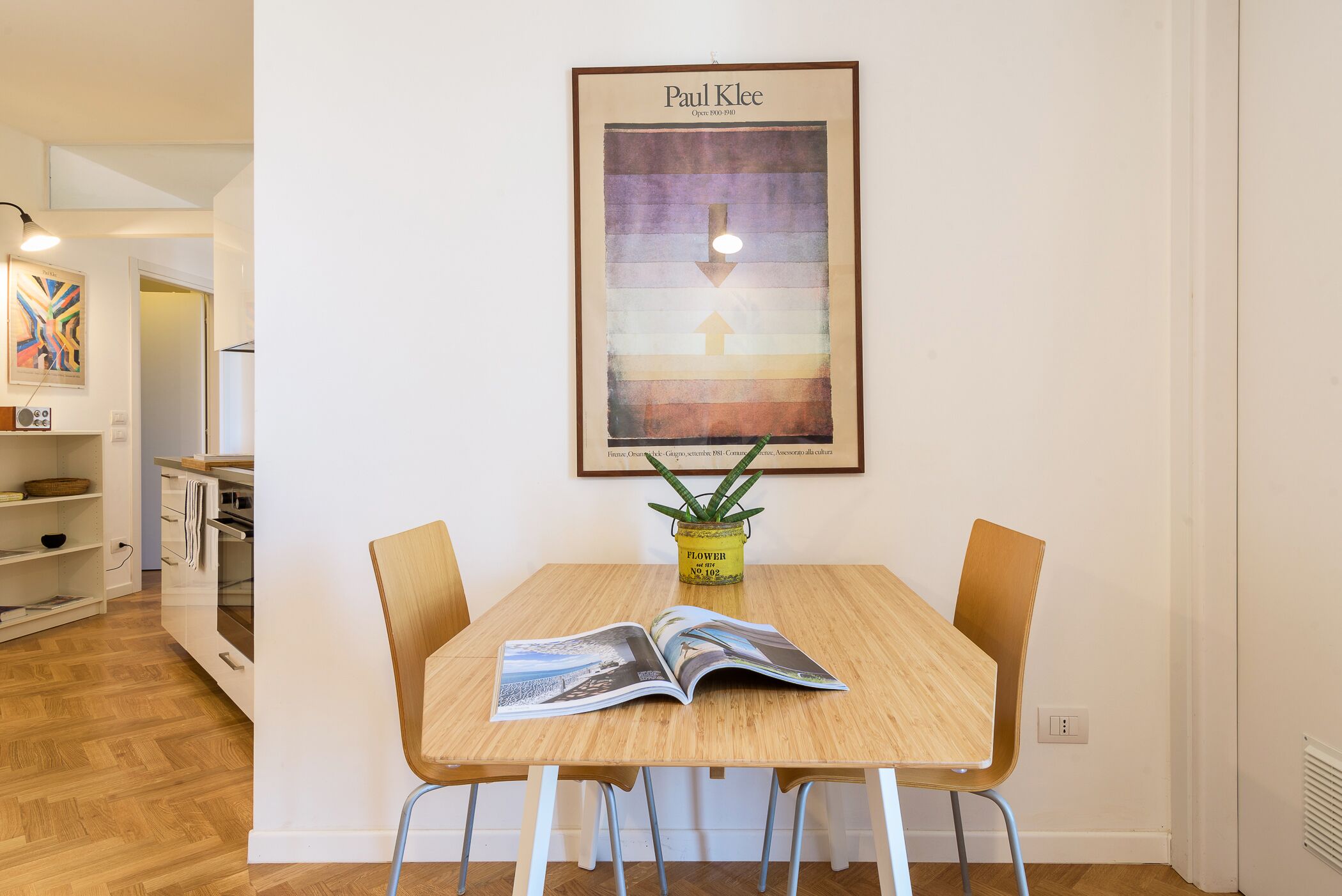 A dining area with a wooden table, two chairs, a framed artwork on the wall, and a small potted plant.