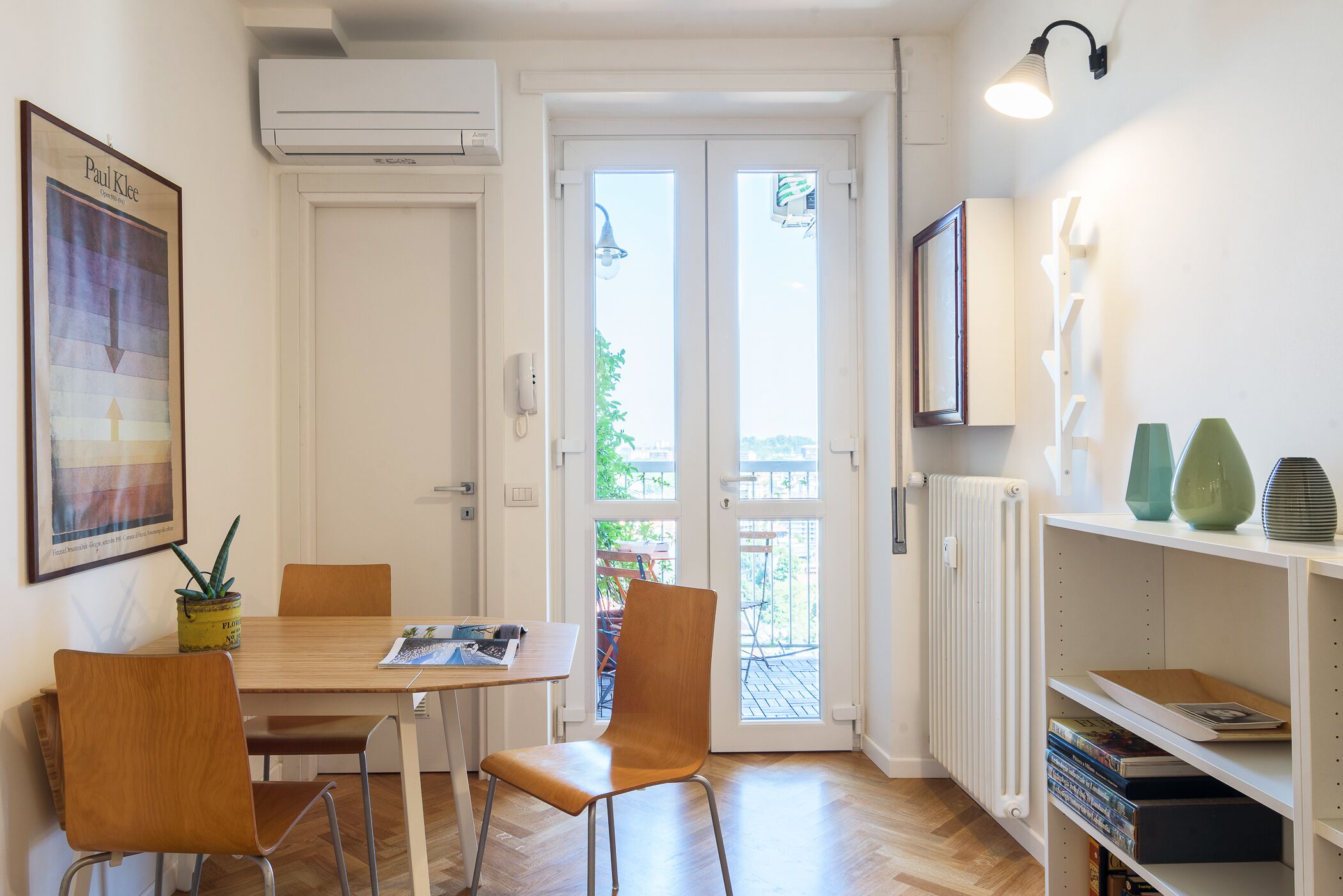 Une pièce comprenant une table à manger en bois, deux chaises, un radiateur, un téléviseur fixé au mur et une bibliothèque contenant des livres et des objets décoratifs.
