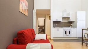A compact living space with a red sofa, a kitchen with white cabinets, and a hallway leading to a bathroom. - JOIVY 1-Bed Flat In Quiet Neighborhood (Milan)