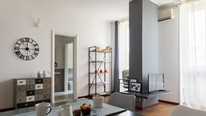 A modern living room with a dining table set for two, a bookshelf, and a large clock on the wall. - JOIVY Superb Flat W/Home Office (Milan)