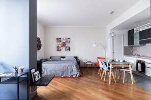 A modern bedroom with a bed, bedside table, and a desk with a chair. There is a small table with chairs in the corner. The room has a wooden floor and a kitchen area with a sink and stove.