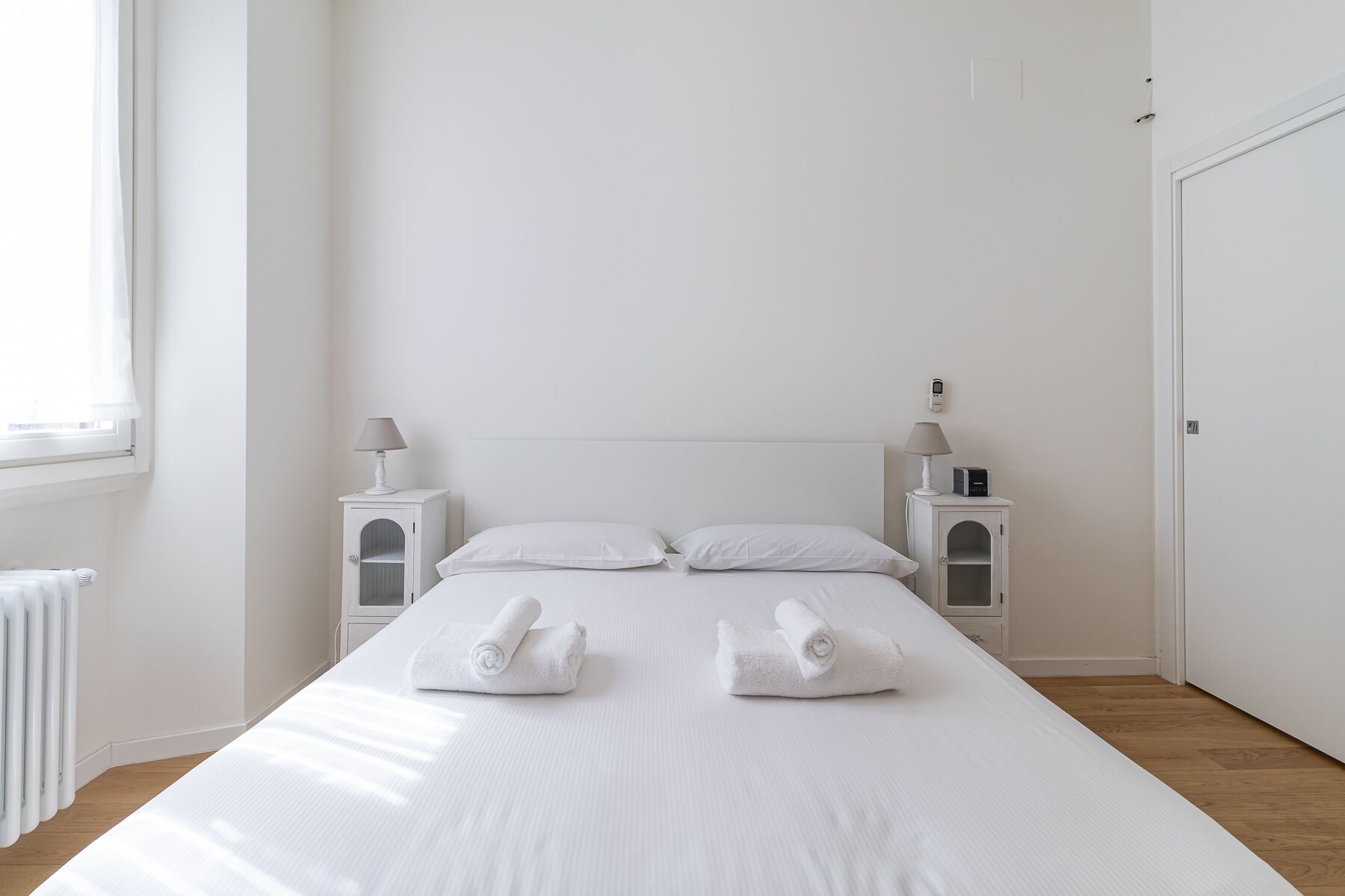 A minimalist hotel room with a large bed, two bedside tables with lamps, a radiator, and a door.