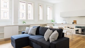 A modern living room with a dark sofa, white cabinets, and a kitchen area with a white countertop and stove. - JOIVY Lovely 1-Bed W/Balcony & Parking (Milan)