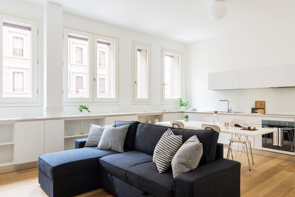 A modern living room with a dark sofa, white cabinets, and a kitchen area with a white countertop and stove.