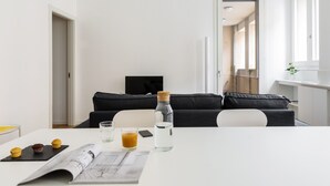 A modern, minimalist living room with a white table, black sofa, and a flat-screen TV. - JOIVY Lovely 1-Bed W/Balcony & Parking (Milan)