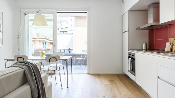 A modern kitchen with white cabinets, a stainless steel oven, and a red backsplash. There is a dining area with a table and chairs, and a sliding glass door leading to a balcony. - JOIVY Stylish 1-Bedroom Flat (Milan)