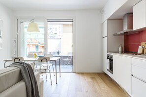 Cozinha moderna com armários brancos, forno de aço inoxidável e painel vermelho. Há uma área de jantar com mesa e cadeiras, e uma porta de vidro deslizante que dá para uma varanda.