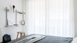 A modern bedroom with a bed, bedside table, and a small stool. There is a shelf with decorative items and a framed picture. - JOIVY Stylish 1-Bedroom Flat (Milan)