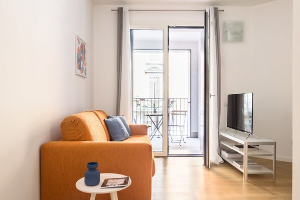 A modern living room with a sofa, a small table, a TV on a stand, and a sliding glass door leading to a balcony.