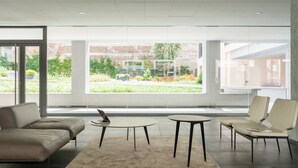 A modern interior with a sofa, two chairs, a small table, and large windows offering a view of greenery and a brick wall.