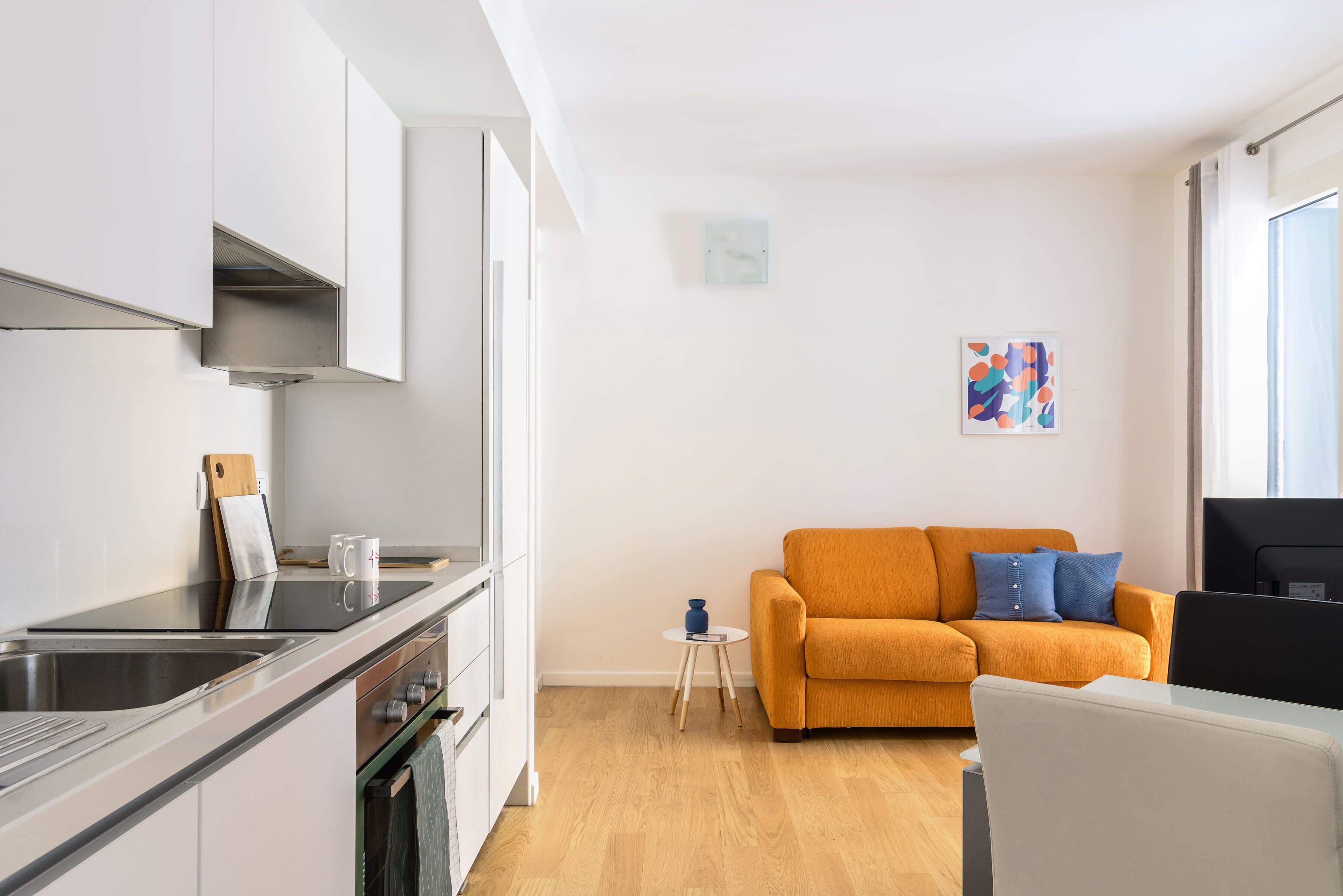 A modern kitchen with a sink, stove, and cabinets, a small side table, and a sofa in a living area with a window.