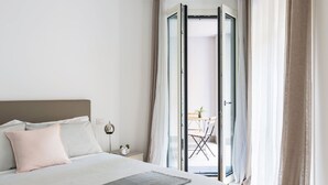 A bedroom with a bed, pillows, a bedside table with a clock, and a view through a glass door to an outdoor area with a table and chairs.