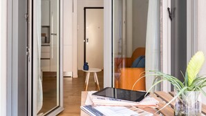 A modern living room with a glass door leading to a kitchen, a wooden table with a tablet and papers, and a potted plant.