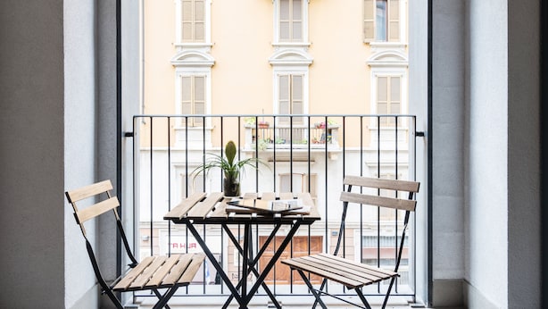 A balcony with a wooden table and chairs, a potted plant, and a view of a building with balconies.