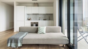 A modern living room with a sofa, a blanket, and a view of a kitchen and balcony.
