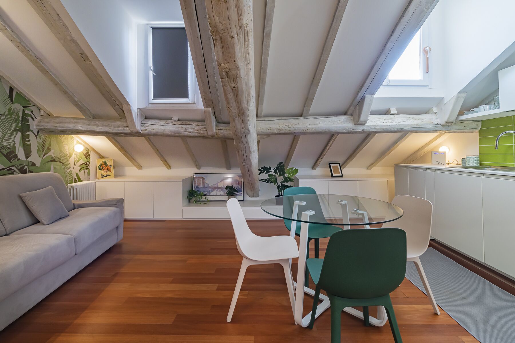 Een knus loft appartement met zichtbare houten balken, een eethoek met een glazen tafel en stoelen, een kitchenette en een woonruimte met een bank en planten.