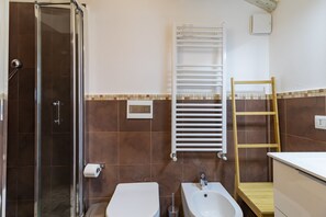Uma casa de banho com cabine de chuveiro em vidro, sanita, bidé, lavatório e barra de toalhas, e uma escada de madeira.