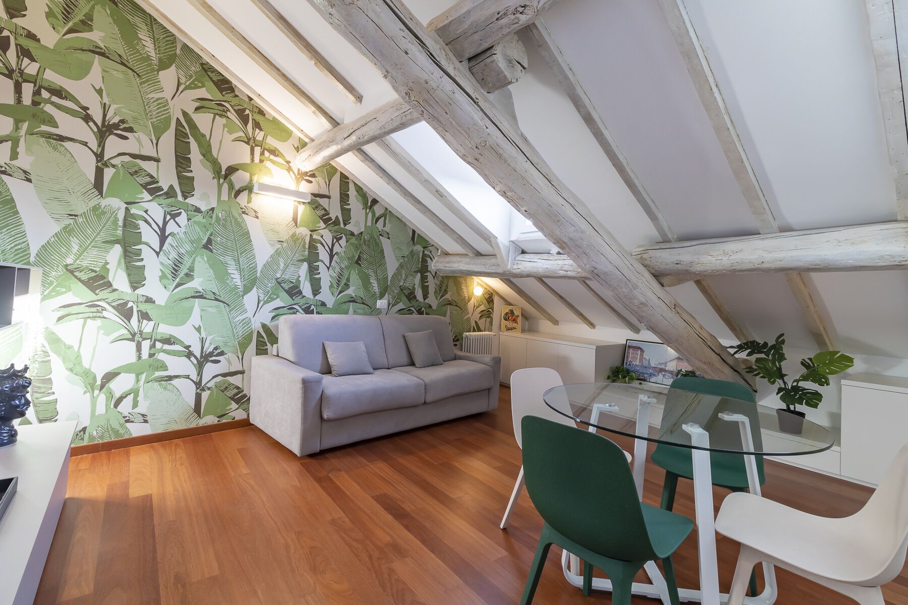 A cozy loft-style room with a wooden floor, a gray sofa, a glass dining table with green chairs, and a wall with a tropical leaf pattern.