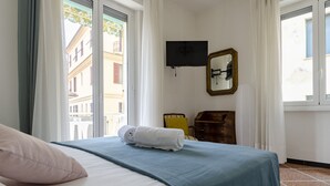 Hotellihuone, jossa on sänky, seinälle asennettu televisio, puinen lipasto ja näkymä rakennuksiin ikkunasta.