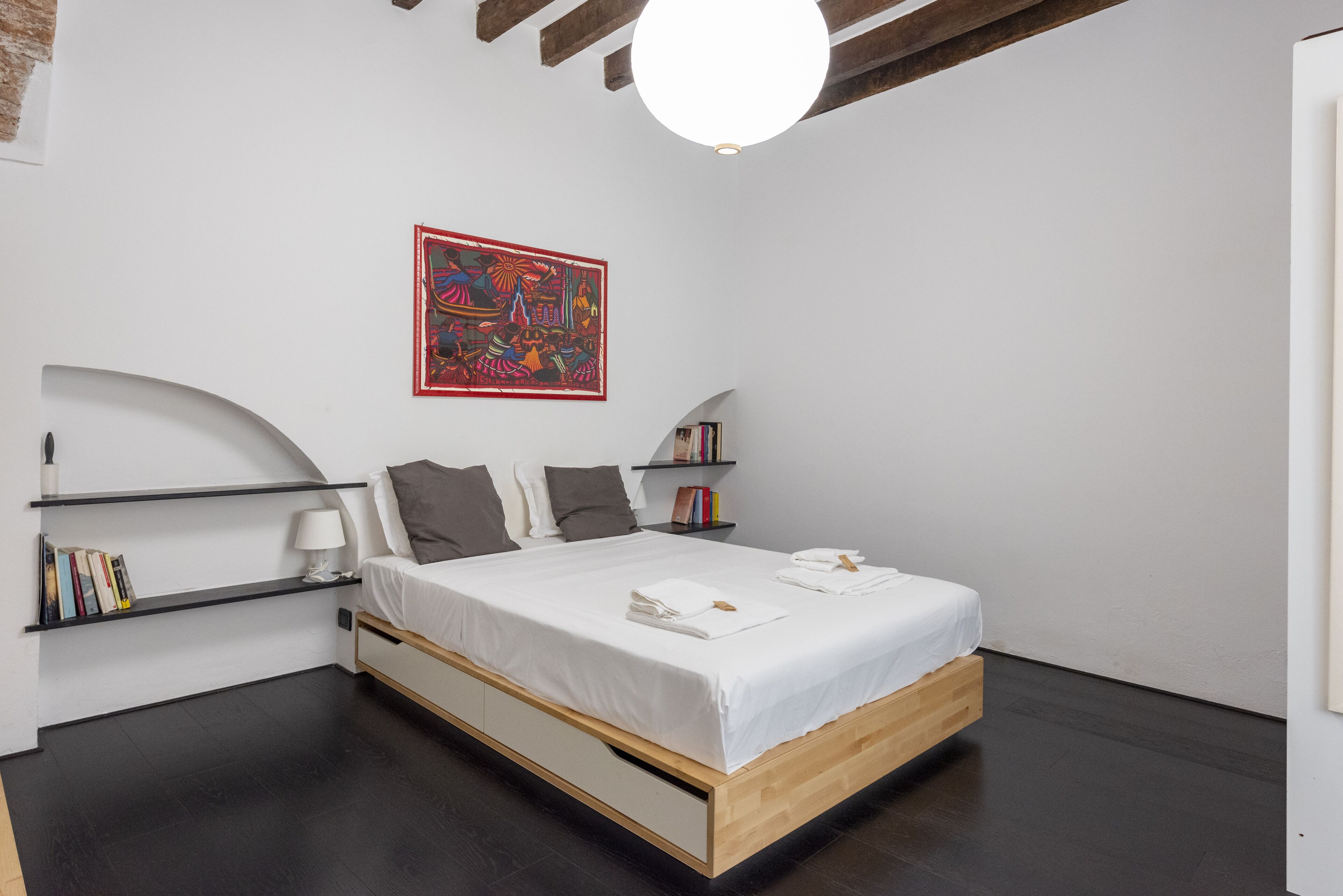 A modern bedroom with a wooden bed, white bedding, a red abstract painting, and built-in shelves with books and a lamp.