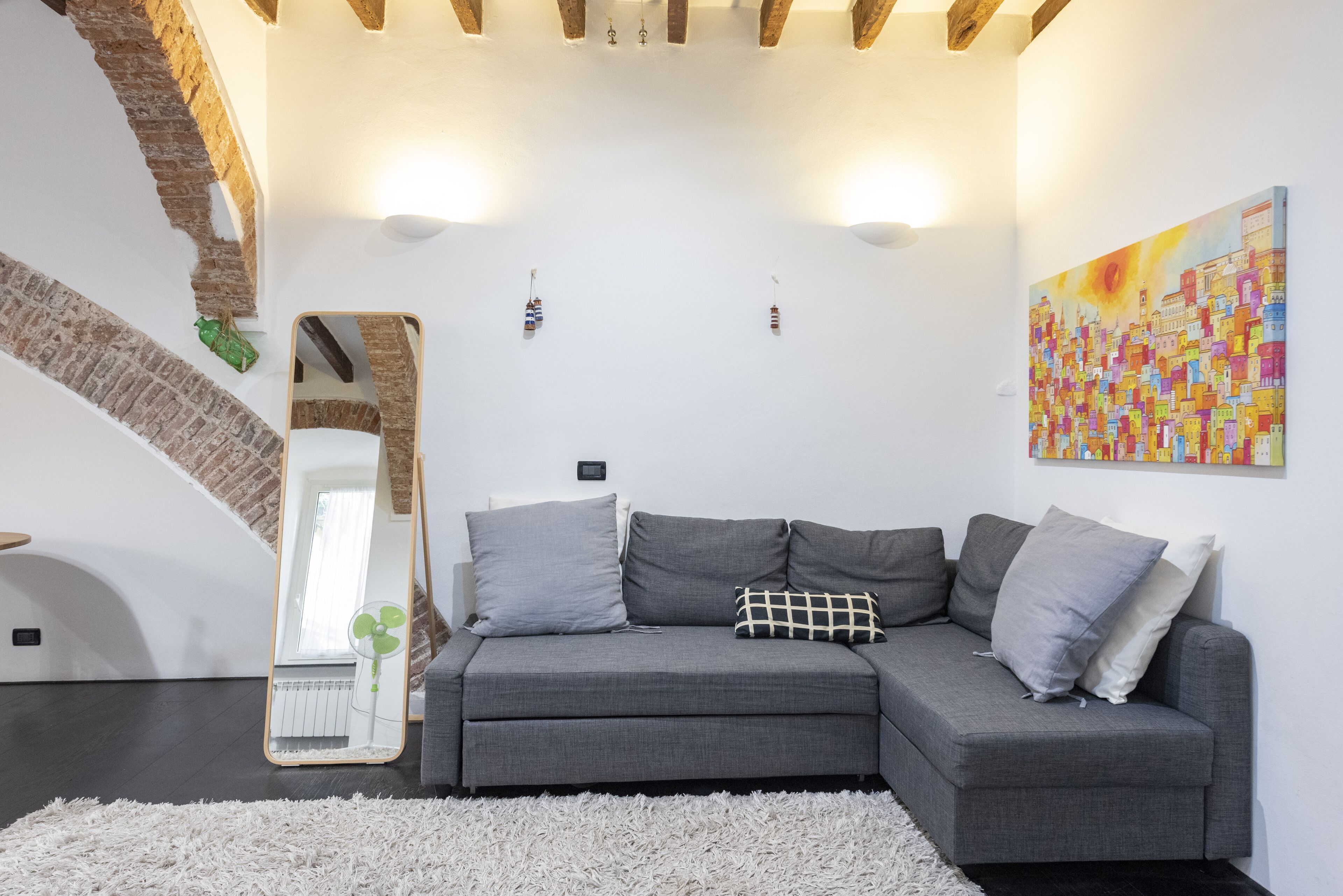 Ein modernes Wohnzimmer mit einem grauen Ecksofa, einem flauschigen weißen Teppich, einem großen Spiegel und einem farbenfrohen abstrakten Gemälde an der Wand.