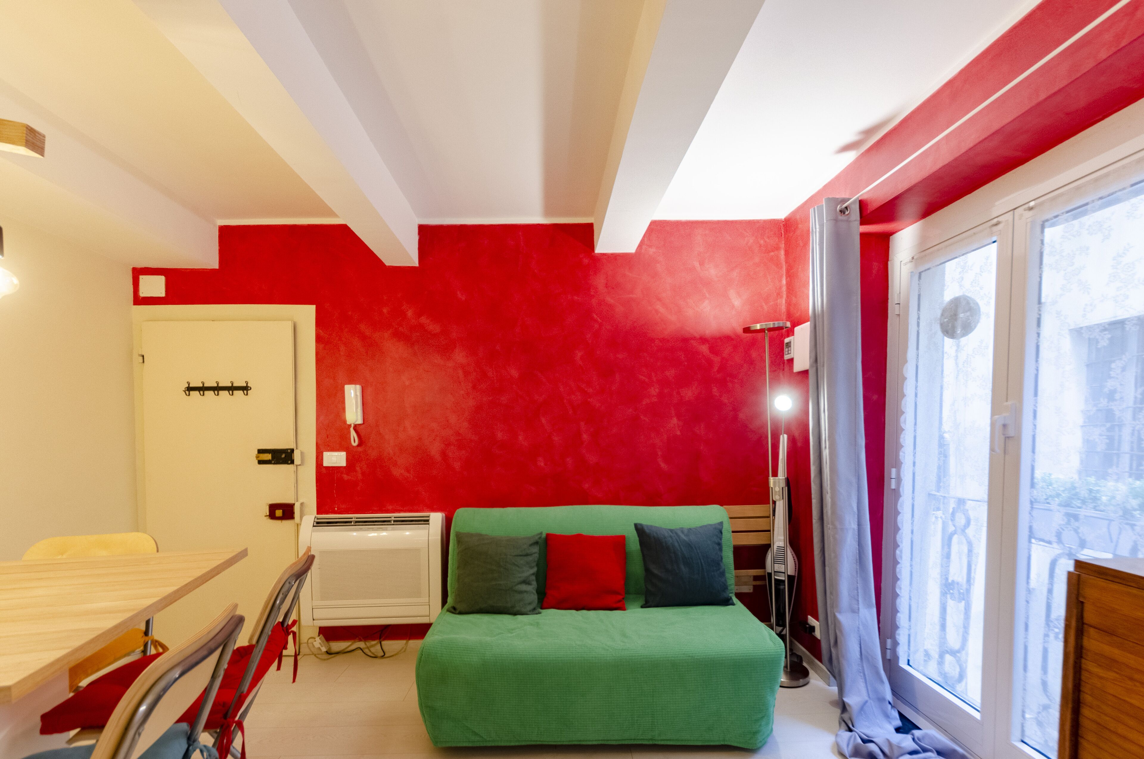A room with a green bed, red walls, a white door, and a window with a view.