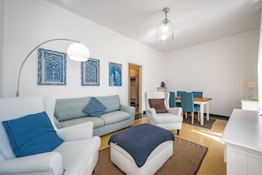 A modern living room with a sofa, armchairs, a coffee table, and a dining area with a table and chairs. There are decorative wall tiles and a ceiling light fixture.