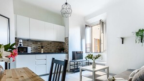 A modern kitchen with white cabinets, a microwave, and a brick backsplash. There is a dining table with chairs, a TV, and a window with a view of the outside. - JOIVY Cosy 1-Bed Flat In Navigli (Milan)