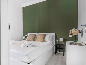 A neatly made bed with white linens and two beige pillows, a small bedside table with a lamp, and a vase with flowers on a dresser. - JOIVY Cosy 1-Bed Flat In Navigli (Milan)