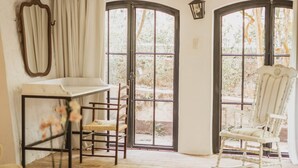 A room with a dining table, two chairs, a rocking chair, a small table with a plant, and large windows with curtains.