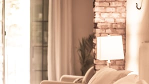 A bed with white linens, a lamp on the bedside table, a brick wall, and a window with curtains.