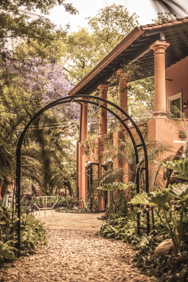 A garden pathway with a metal archway, surrounded by lush greenery and a building with columns.