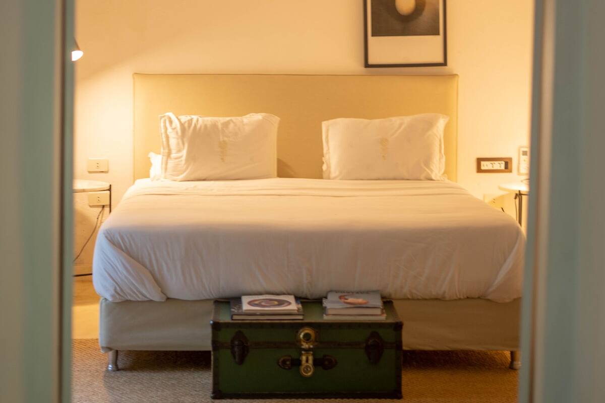 Une chambre à coucher avec un lit, des oreillers, une table de chevet, une commode, un tableau encadré au mur et une valise verte posée sur le sol.
