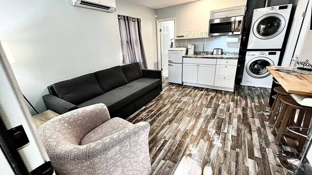 Ett kompakt bostadsutrymme med en soffa, en mönstrad fåtölj, en liten köksdel och en tvättmaskin.