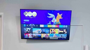 En platt-TV är monterad på väggen och visar ett gränssnitt för en streamingtjänst med olika innehållsalternativ. Under TV:n finns en bardisk i trä med två barstolar.