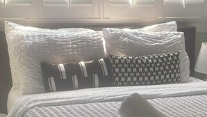 Ein ordentlich bezogenes Bett mit weißer Bettwäsche und gemusterten Kissen, ein aufgerolltes Handtuch auf dem Bett sowie ein Fenster mit Jalousien im Hintergrund.
