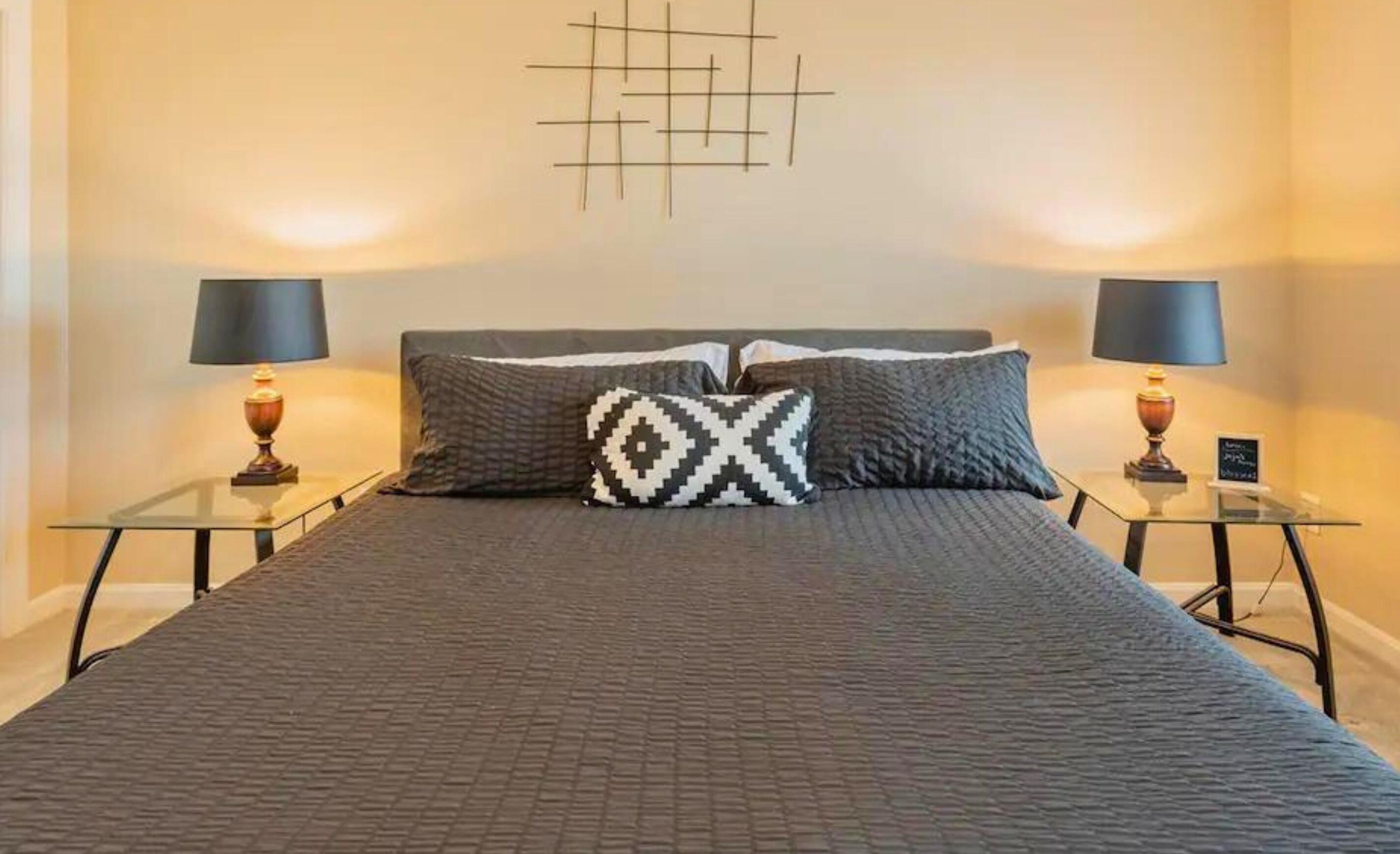 Una cama bien hecha con colcha gris y dos almohadones estampados, flanqueada por dos mesitas de noche con lámparas y una obra de arte en la pared.